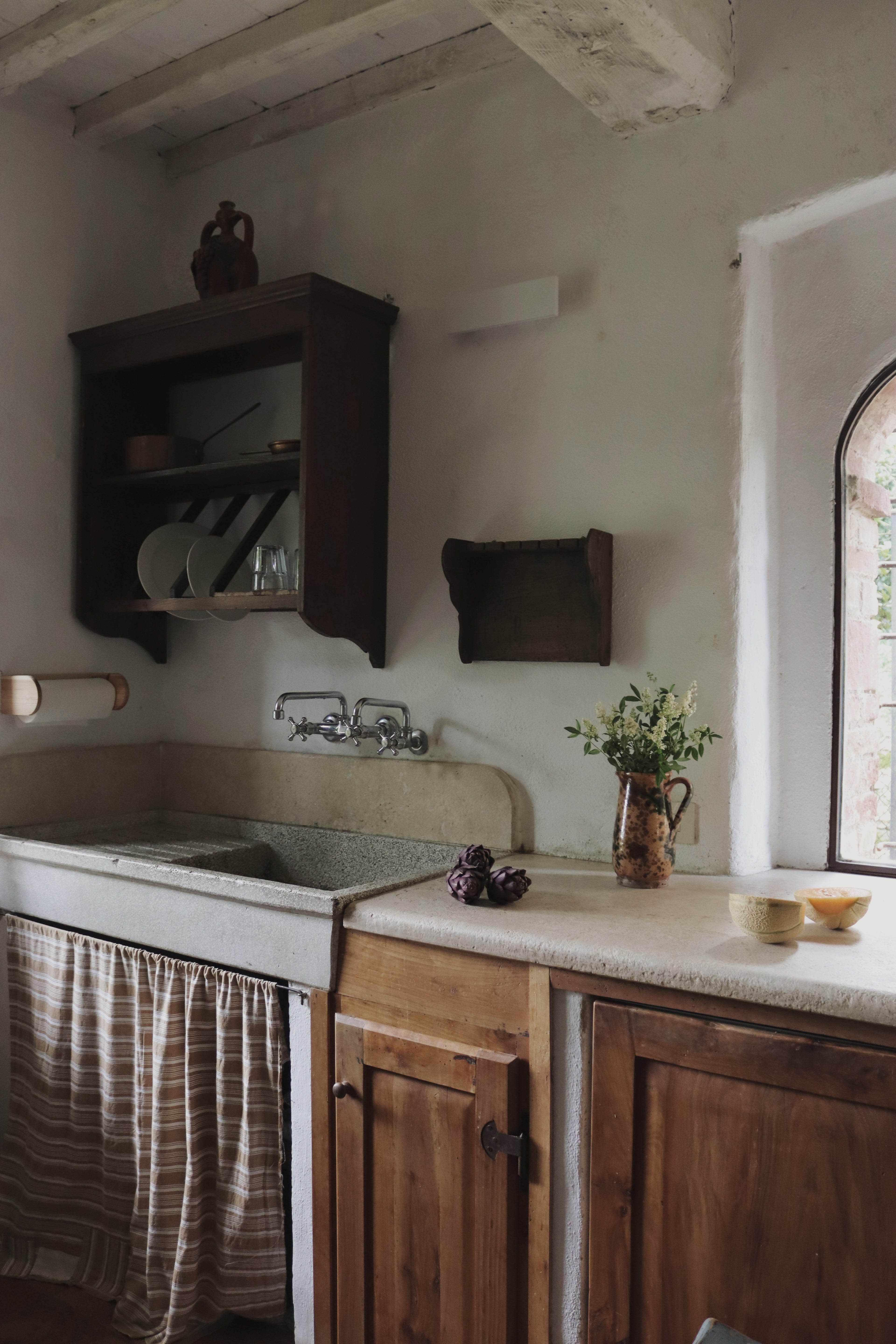 Rustic Tuscan farmhouse kitchen with stone sink, aged wood cabinets, artichokes, and wildflowers in a ceramic pitcher