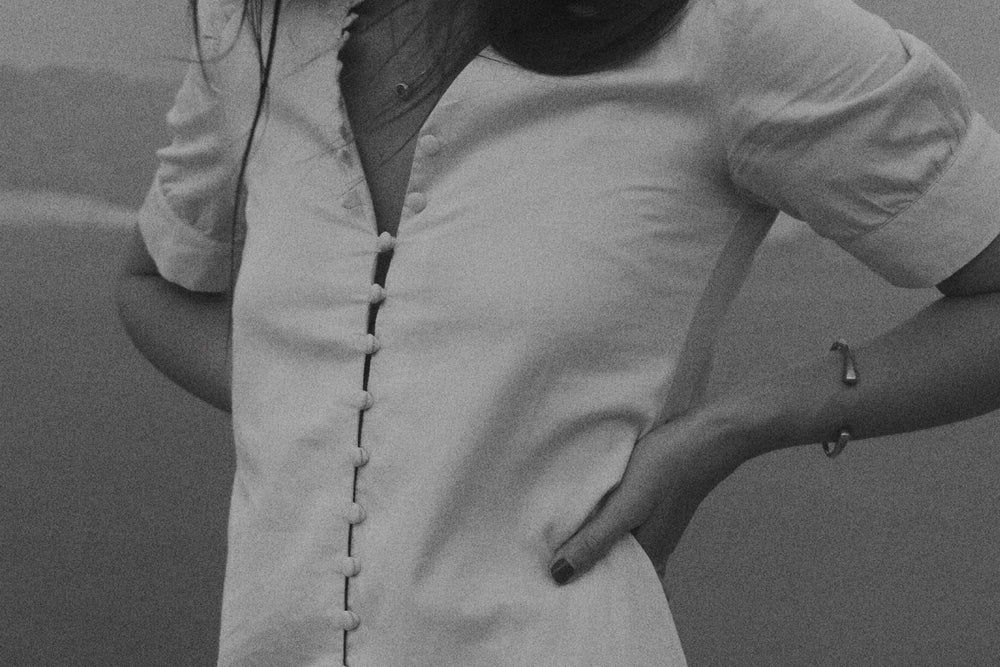 A woman wearing a short-sleeve white linen dress with a front button closure, standing outdoors on a rocky surface with a blurred natural background.