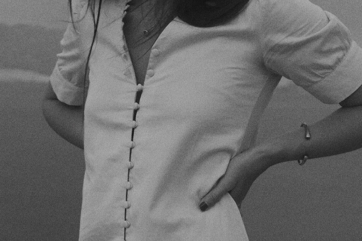 A woman wearing a short-sleeve white linen dress with a front button closure, standing outdoors on a rocky surface with a blurred natural background.