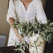 Woman in The Lydia cotton-bamboo dress arranging wild olive branches in a ceramic vessel on a dark wood table