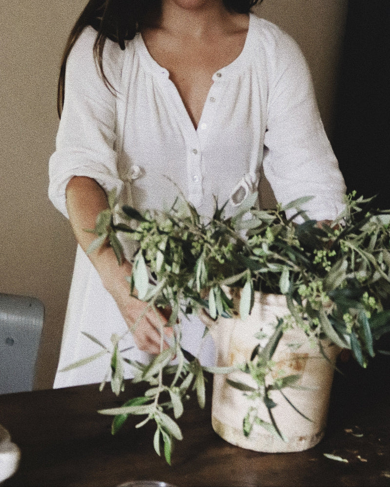 Woman in The Lydia cotton-bamboo dress arranging wild olive branches in a ceramic vessel on a dark wood table