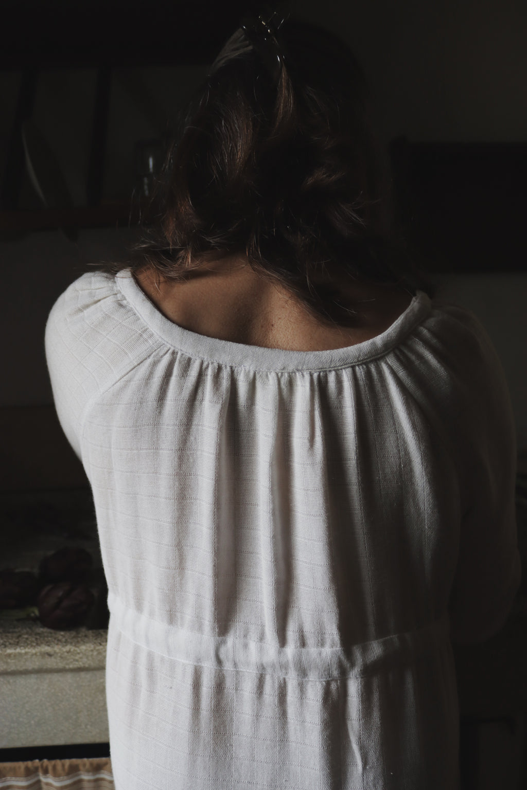 Back view of The Lydia cotton-bamboo dress showing gathered scoop neckline and textured fabric, dim Tuscan interior