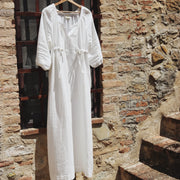 The Lydia cotton-bamboo dress hanging on a wooden hanger against a sun-drenched Tuscan stone wall