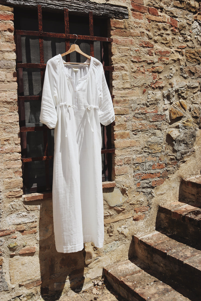 The Lydia cotton-bamboo dress hanging on a wooden hanger against a sun-drenched Tuscan stone wall