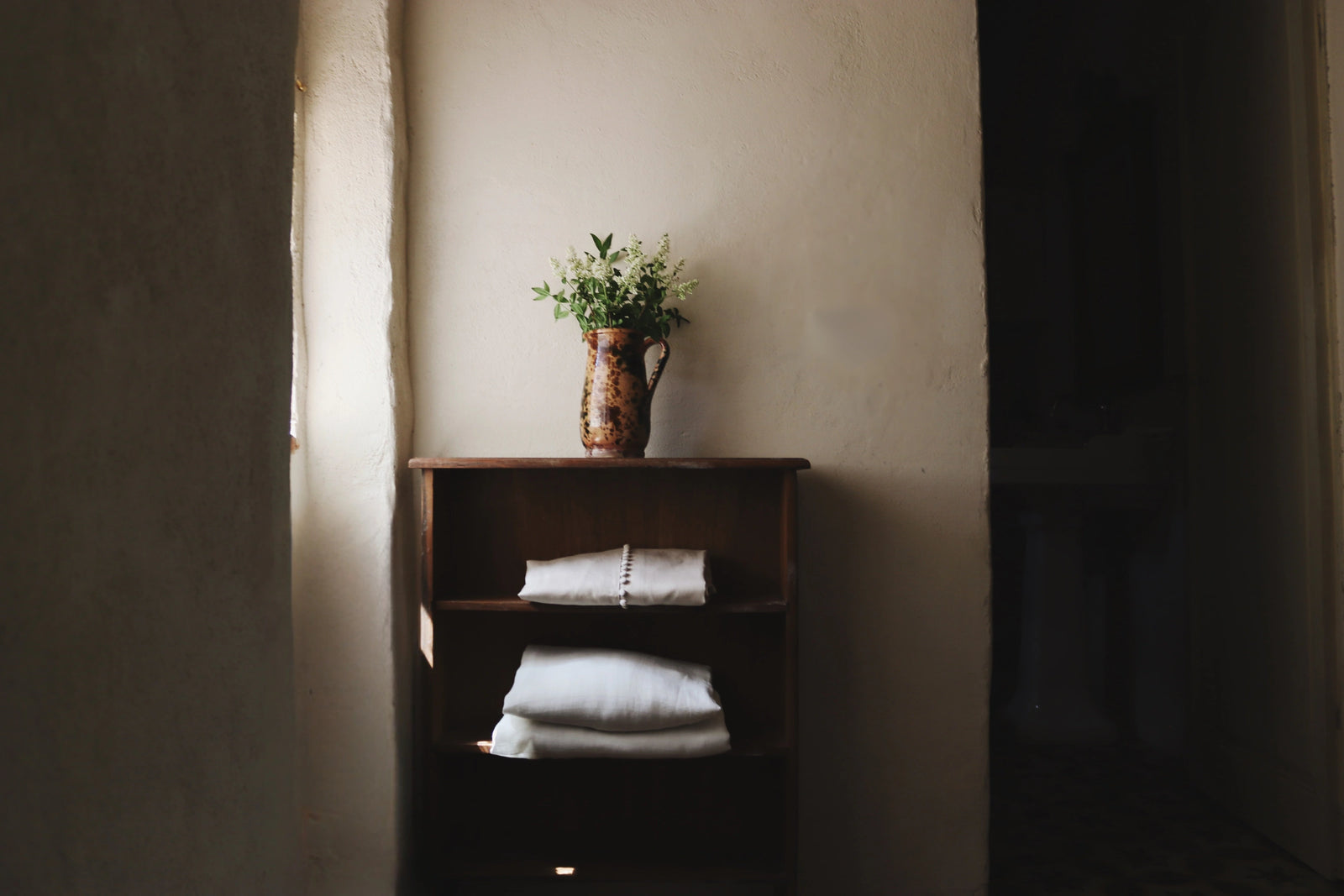 Folded white linen dresses on a worn wooden shelf beside a ceramic pitcher with wildflowers, Tuscan stone alcove