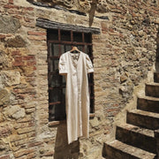 The Thelma cotton dress in cream hanging against a sun-lit Tuscan stone wall with iron window and stone steps