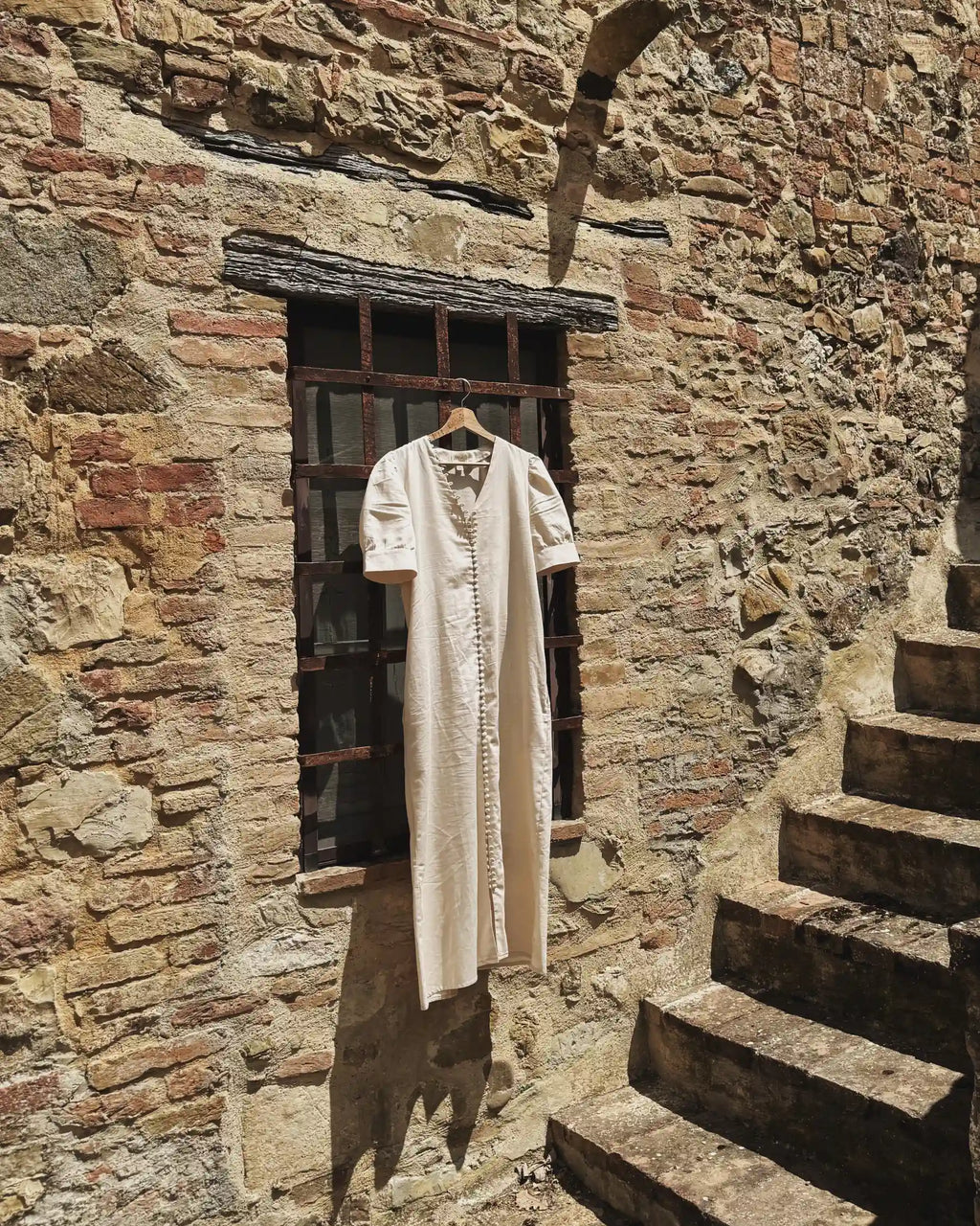 The Thelma cotton dress in cream hanging against a sun-lit Tuscan stone wall with iron window and stone steps