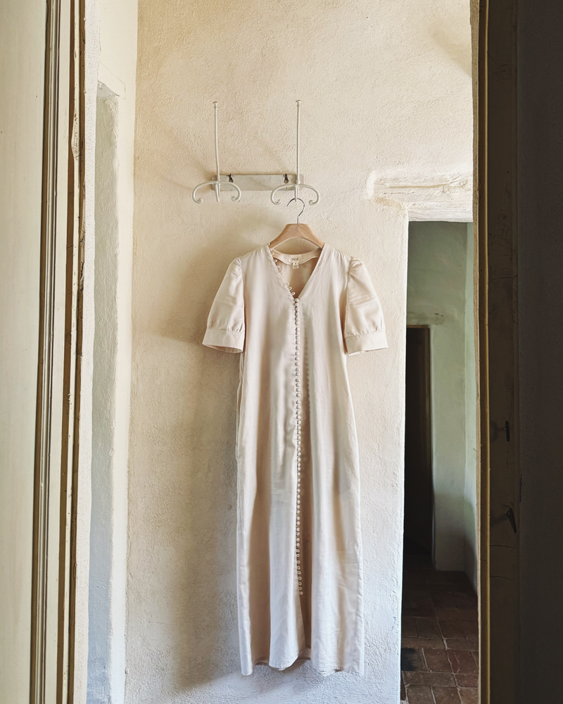 The Thelma cotton dress in cream hanging on an iron hook against a plastered Tuscan wall, full length view